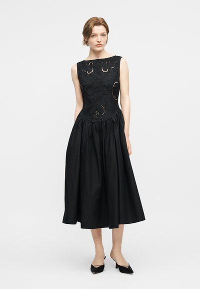 Woman with short hair wears sleeveless black midi dress with embroidered bodice and gathered skirt, paired with black pointed heels.