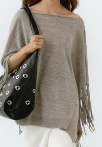 Hellgrauer übergroßer Pullover mit weitem Ausschnitt und Fransenverzierungen an den Ärmeln. Schwarze Tasche mit silbernen Ösenakzenten.