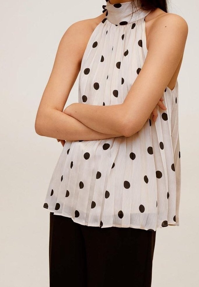 Femme portant un haut dos nu blanc à pois noirs et un pantalon noir, les bras croisés sur la poitrine.