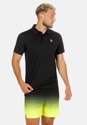 Man die een zwart poloshirt en geel-naar-zwart verlopende korte broek draagt, staat en kijkt naar rechts.