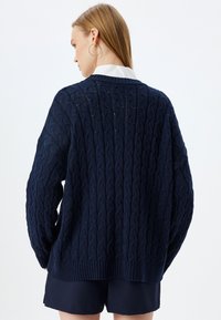 Maglione navy a trecce con una vestibilità rilassata, polsini e orlo a coste, caratterizzato da motivi testurizzati. Indossato sopra una camicia bianca con colletto e pantaloni shorts.
