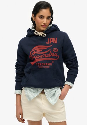 Donkerblauwe hoodie met een voorzak in kangoeroestijl, voorzien van rode grafische tekst en logo. Gedragen over een licht gestreept shirt en gecombineerd met witte shorts.