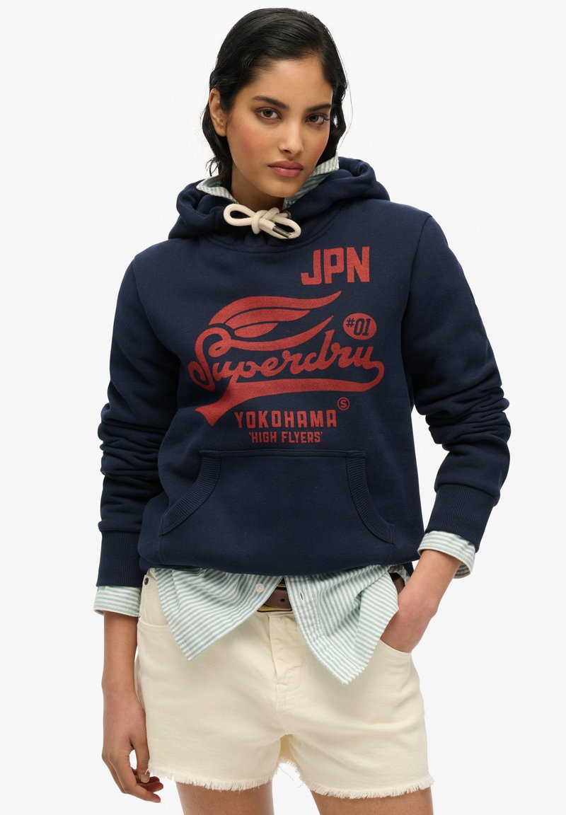 Sweat à capuche bleu marine avec une poche kangourou à l'avant, arborant un texte graphique rouge et un logo. Porté par-dessus une chemise à rayures claires et associé à un short blanc.