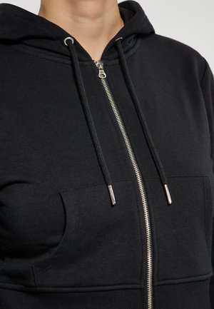 Czarna bluza z kapturem na zamek błyskawiczny, związywana, z teksturowanej tkaniny, z kieszeniami z przodu, srebrnym okuciem i dopasowanym krojem. Zbliżenie na obszar klatki piersiowej.