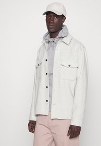 Veste en flanelle gris clair avec des boutons noirs, deux poches poitrine, superposée sur un sweat à capuche gris, accompagnée de pantalons rose clair.