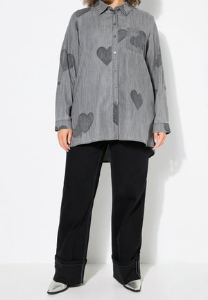 Kobieta w za dużej szarej koszuli na guziki z ciemnymi wzorami w kształcie serc, czarne szerokie spodnie i srebrne spiczaste buty.