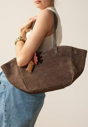 Femme en haut sans manches et jean tenant un grand sac fourre-tout en daim marron avec un porte-clés floral sur l'épaule, sur un fond clair.