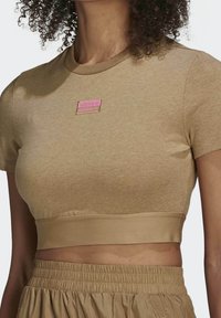 T-shirt corta color sabbia in misto cotone, con scollatura rotonda e un grande patch del logo Adidas rosa sul petto. Texture liscia.