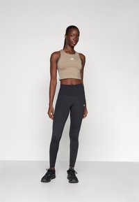 Tan avkortet tanktop med New Balance-logo, kombinert med svarte høyt midjepants. Modellen har på seg svarte treningssko og står foran en ensfarget bakgrunn.