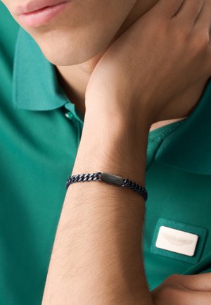 Uomo con polo verde con distintivo, che si appoggia la mano sul collo, mostrando un braccialetto a catena nero con chiusura rettangolare al polso.