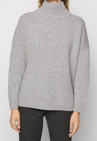 Grå rullekravesweater med ribbet krave og løs pasform, der har en blød tekstur og lange ærmer, kombineret med mørke bukser.