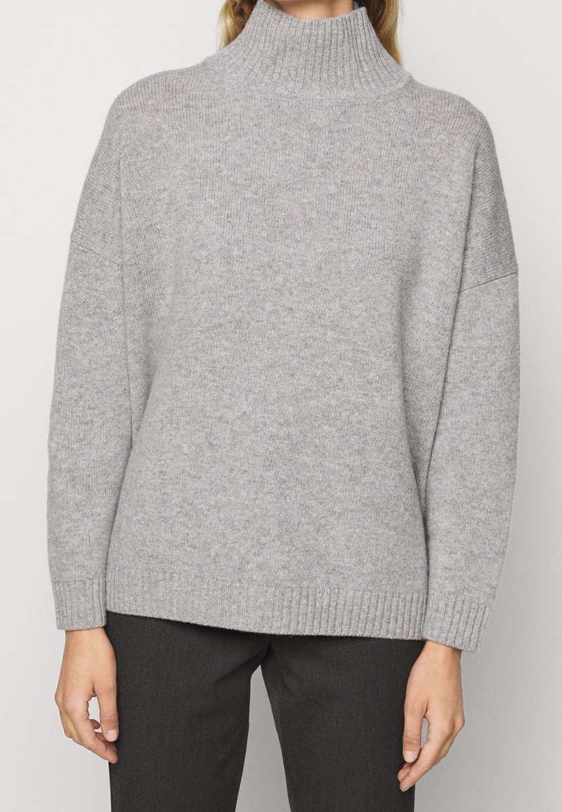 Grå rullekravesweater med ribbet krave og løs pasform, der har en blød tekstur og lange ærmer, kombineret med mørke bukser.