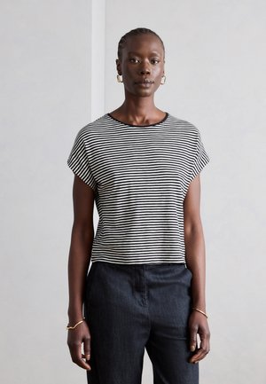 Kvinde med fletninger iført sort-hvid stribet kortærmet skjorte, sorte bukser, guldringeørede øreringe og armbånd, står mod en hvid væg.