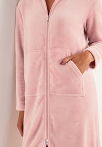 Roze fleece badjas met ritssluiting, lange mouwen en twee voorzakken. Zachte textuur en zichtbare stiksels langs de naden.