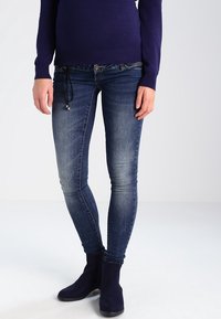 Kapehtav jeansit keskmiselt sinises denimkangas, millel on kulumis- ja venitusjooned. Kanda tumesinise sviitriga ja mustade pahkluu saapadega. Reguleeritav nöörivöö.