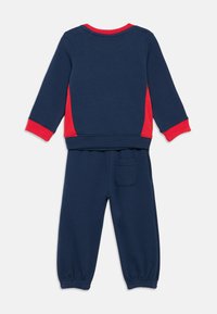 Mørkeblå og rød langærmet sweatshirt til tumlinge samt matchende bukser med elastiske manchetter og en baglomme på ensfarvet baggrund.