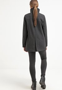 Dunkelgrauer Mantel aus Wollmischung mit hohem Kragen, seitlichen Taschen und einem mittigen Rückenschlitz. Kombiniert mit schwarzen Leggings und Stiefeletten.