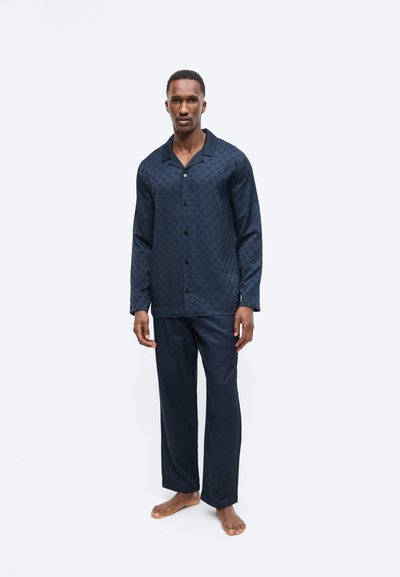 Férfi áll mezítláb, sötétkék, hosszú ujjú, gombolós ingben és hozzáillő, bő nadrágban, egyszerű fehér háttér előtt.