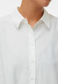 Chemise à col blanc avec un bouton à l'avant, ornée de petits strass décoratifs répartis uniformément sur le tissu pour un détail supplémentaire.