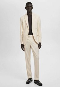 Helle beige Anzugjacke und Hose, kombiniert mit einem schwarzen Hemd. Der Anzug hat eine taillierte Passform und eine glatte Textur, mit einem Ein-Knopf-Verschluss.