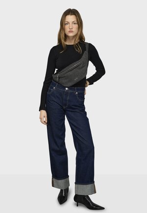 Mujer vestida con una blusa negra de manga larga, jeans oscuros con los bajos doblados, botines negros y un bolso gris cruzado sobre el cuerpo, posando frente a un fondo liso.