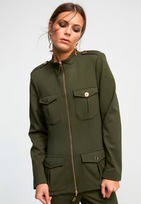 Veste verte olive en tissu structuré, dot ée d'un col montant, de zips dorés et de deux poches avant avec fermetures à bouton.