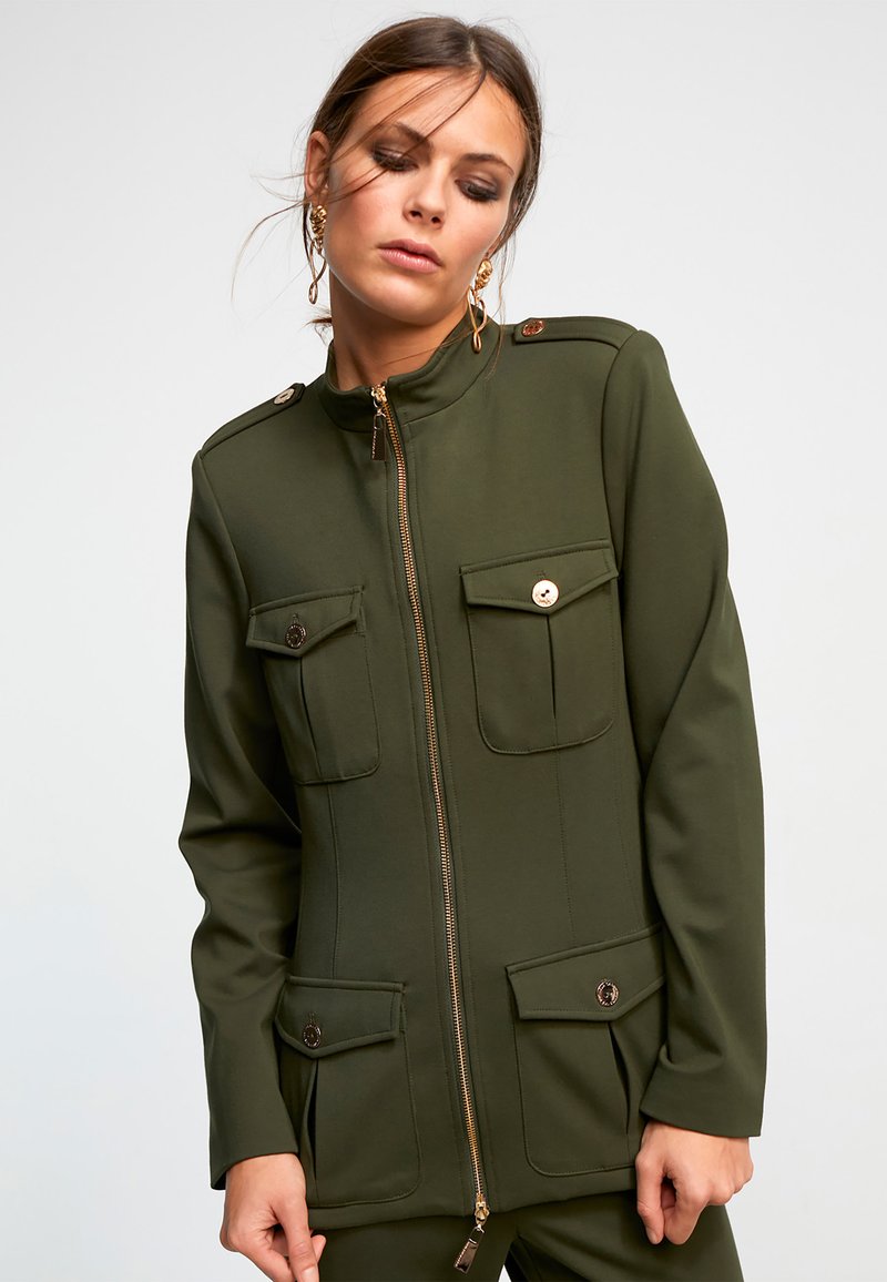 Veste verte olive en tissu structuré, dotée d'un col montant, de zips dorés et de deux poches avant avec fermetures à bouton.