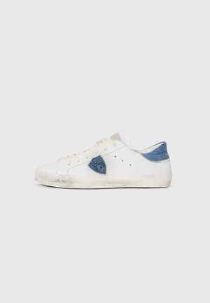 Zapatillas bajas blancas con un acento en el talón de gamuza azul y un parche de tela azul en el costado. Con suela de goma texturizada y cordones.