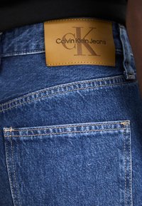Plave traperice s smešnim kožnim oznakama utisnutim s "Calvin Klein Jeans" i stilizovanim "CK" logom. Vidljivi detalji šivanja.