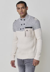 Jersey de punto con cuello alto, en colores gris y crema, con textura acanalada, bolsillos en el pecho y detalles de botones en los hombros.