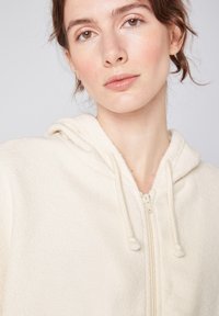 Haut à capuche en polaire couleur crème avec une fermeture éclair à l'avant, une capuche à cordon et une texture douce. Design minimaliste avec une coupe décontractée et des détails subtils.