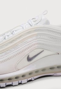 Gros plan d'une basket blanche Nike avec des matériaux en mesh et en cuir, des coutures visibles, des passants pour lacets, et un détail de semelle à coussin d'air transparent.