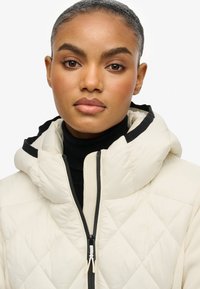 Kurtka puchowa w kolorze off-white, z diamentowym pikowaniem, czarnymi akcentami, wysokim kołnierzem i zamkiem błyskawicznym z przodu. Gładka tekstura i lekka.