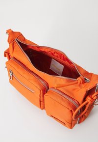 Pomarańczowa nylonowa torebka na ramię z dwiema zapinanymi na zamek przednimi kieszeniami, pojemną komorą główną oraz widoczną metką marki wewnątrz.