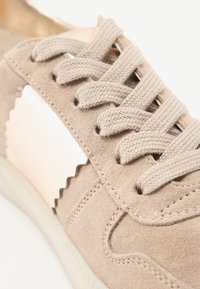 Beige semsket sneakers med strukturert søm og en bølget hvit dekorasjon. Har flate beige snører og en gummisåle, nærbilde.
