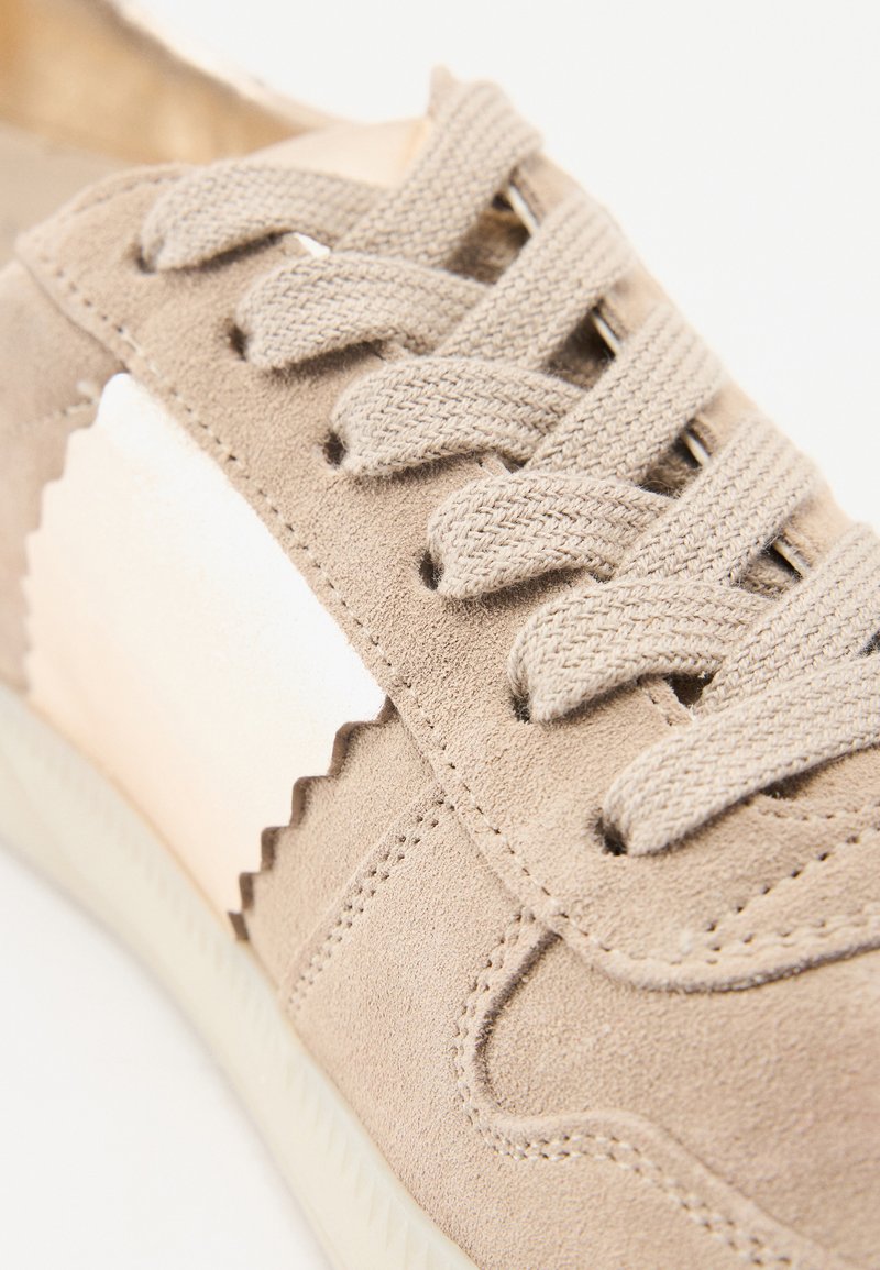 Sneaker in suede beige con cuciture testurizzate e un accento bianco a onde. Presenta lacci piatti beige e una suola in gomma, visione ravvicinata.