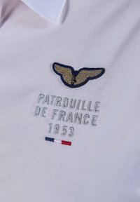 Tissu blanc avec des ailes dorées brodées et le texte argenté "Patrouille de France 1953" au-dessus d'une petite bande bleue, blanche et rouge.