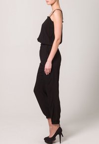 Combinaison noire à bretelles spaghetti, haut ample, taille élastique, jambes fuselées et poignets, associée à des chaussures à talons hauts noires.