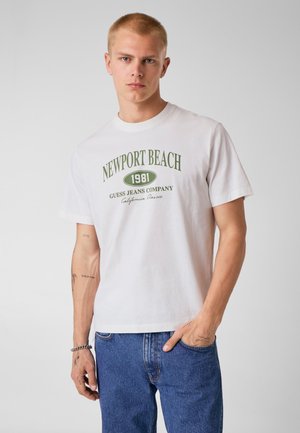 Jeune homme aux cheveux blonds courts portant un t-shirt blanc Newport Beach 1981 et un jean bleu, debout avec une main dans la poche.