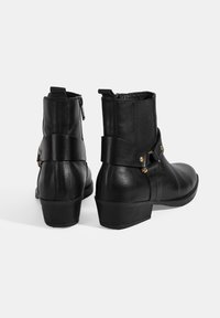 Bottines en cuir noir avec un bout arrondi, fermeture éclair, panneaux latéraux élastiques et une sangle décorative avec des éléments en métal doré.