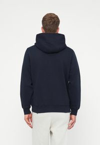 Uomo che indossa un felpa con cappuccio blu navy e pantaloni grigi chiari, in piedi con la schiena rivolta verso la fotocamera su uno sfondo bianco semplice.