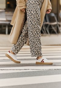 White sneakers with red accents and brown soles, paired with beige pants featuring a black leopard print, walking on a striped crosswalk.