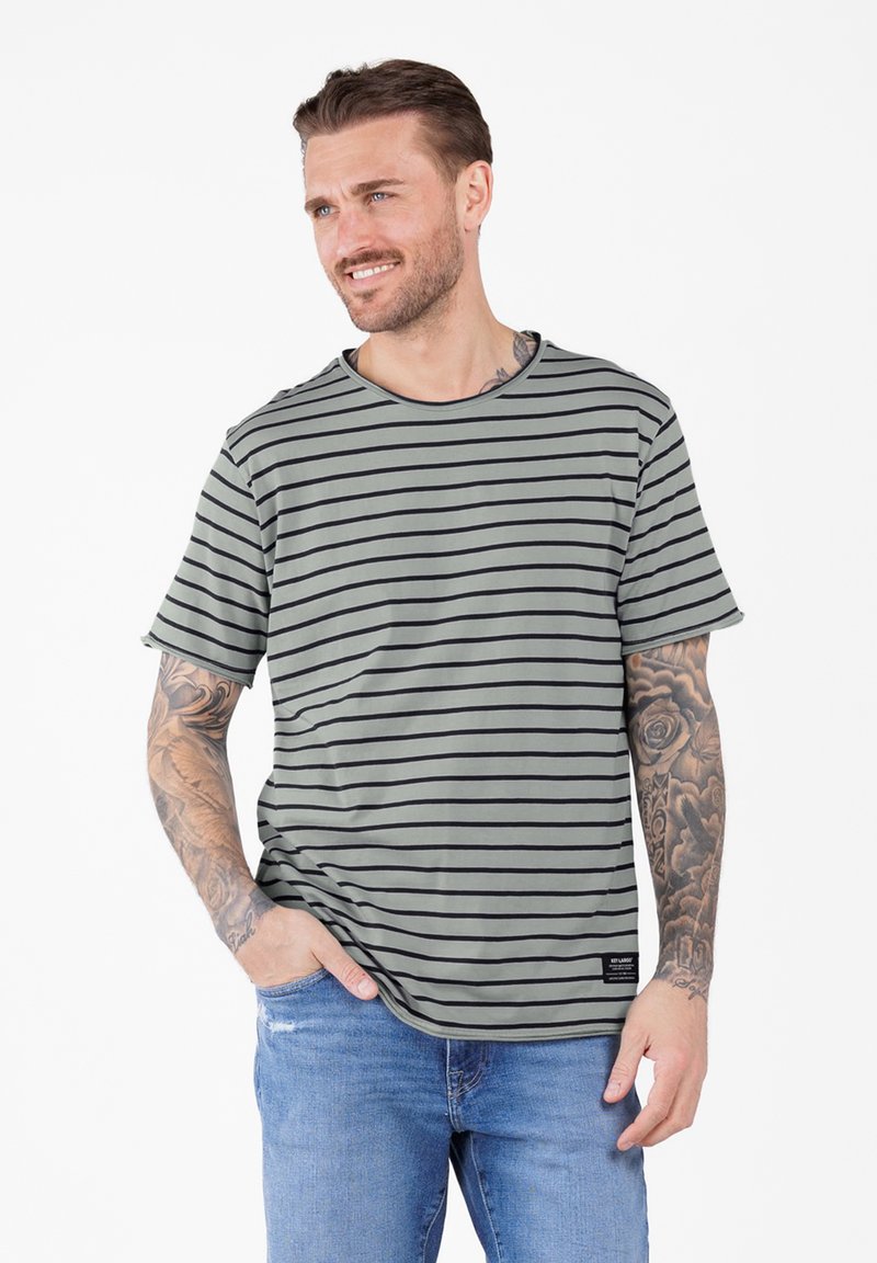 Hombre con tatuajes que lleva una camiseta a rayas gris y negra y jeans azules, sonriendo y mirando hacia un lado con una mano en el bolsillo.