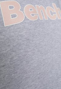Tessuto grigio chiaro con grandi lettere a pois rosa e bianche che formano la parola "Bench" in carattere bold.