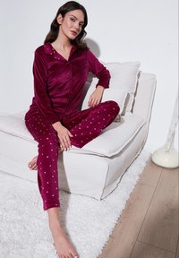Burgundska velur pidžama s pregačom s gumbima i hlačama s točkicama, s malim uzorcima srca. Model sjedi na svijetloj sofi.