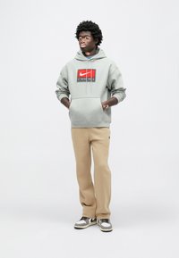 Γκρι φούτερ με κουκούλα της Nike με κόκκινο και μαύρο λογότυπο, συνδυασμένο με μπεζ παντελόνι και γκρι αθλητικά παπούτσια. Το μοντέλο στέκεται με τα χέρια στις τσέπες μπροστά σε ένα φωτεινό φόντο.