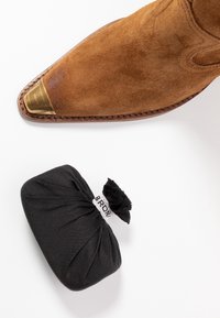 Botas de vaquero de ante marrón con punta de metal al lado de un estuche aplicador de betún negro sobre una superficie blanca.