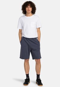 White cotton T-shirt with a pocket, paired with navy blue shorts and black sneakers. Model has curly hair and stands with hands in pockets.