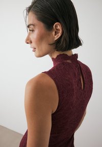 Burgundy spetsklänning med hög hals, vågig trim och nyckelhål på baksidan; pryds av intrikata blommönster och strukturerad tyg.