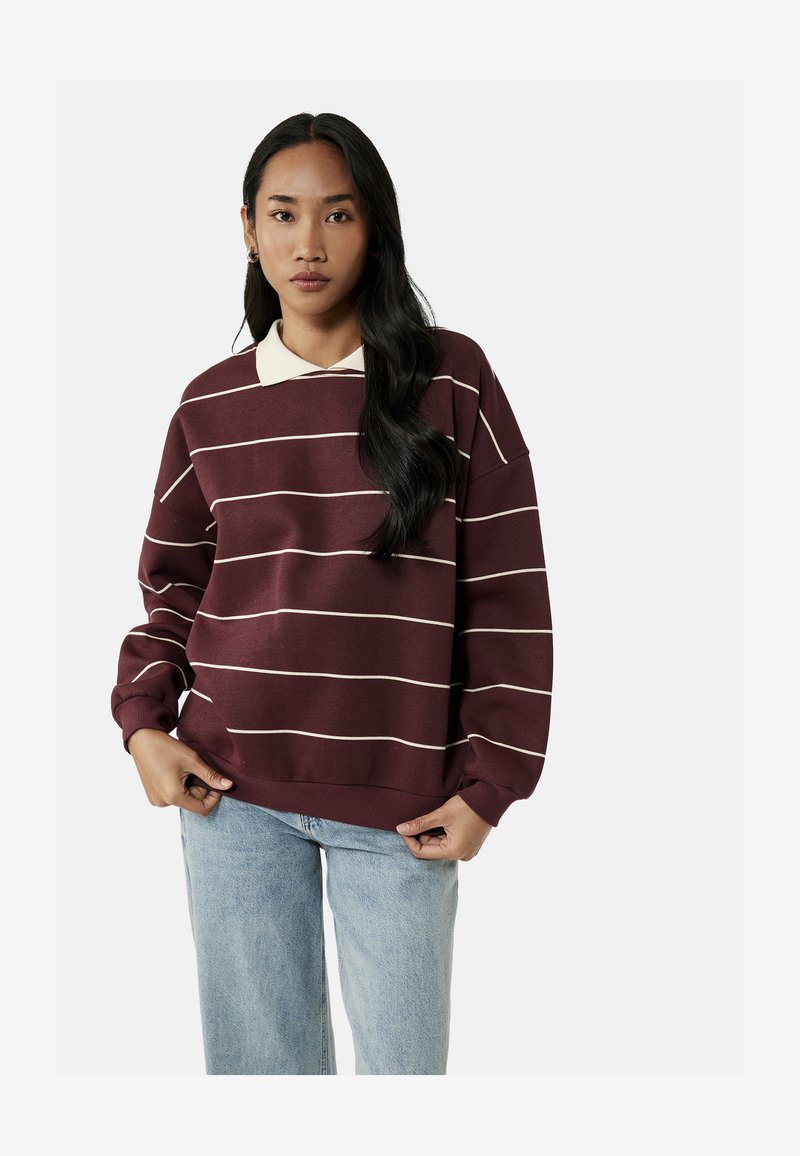Femme aux longs cheveux noirs portant un pull rayé bordeaux par-dessus une chemise blanche à col et un jean bleu clair, debout devant un fond uni.